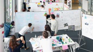 Zu sehen ist ein Raum der HPI School of Design Thinking mit mehreren Studenten und White-Boards, wo die Studierenden dran Arbeiten.