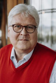 Älterer Mann mit weißen, kurzen Haaren, einer dunklen Brille und einem roten, ärmellosen Pullover