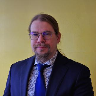 Portrait eines Mannes mit runder Brille und Bart, der ein dunkelblaues Chakett sowie Krawatte trägt.